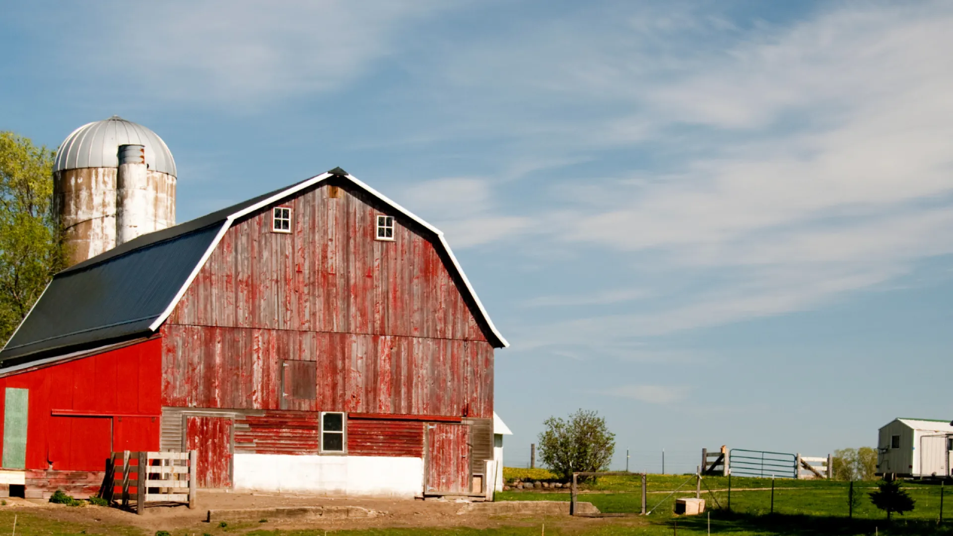 Red weathered barn with silver silo and clear blue sky on a rural farm landscape with green trees.