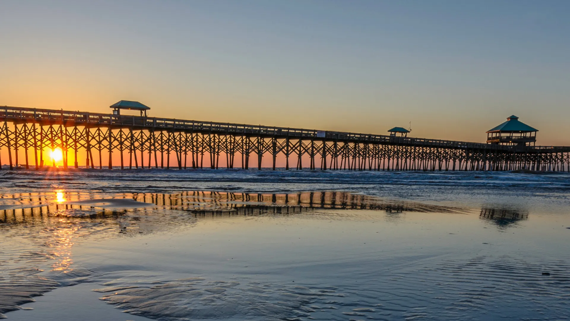 Sunset over a long wooden pier with reflections on wet sandy beach and calm ocean in the background