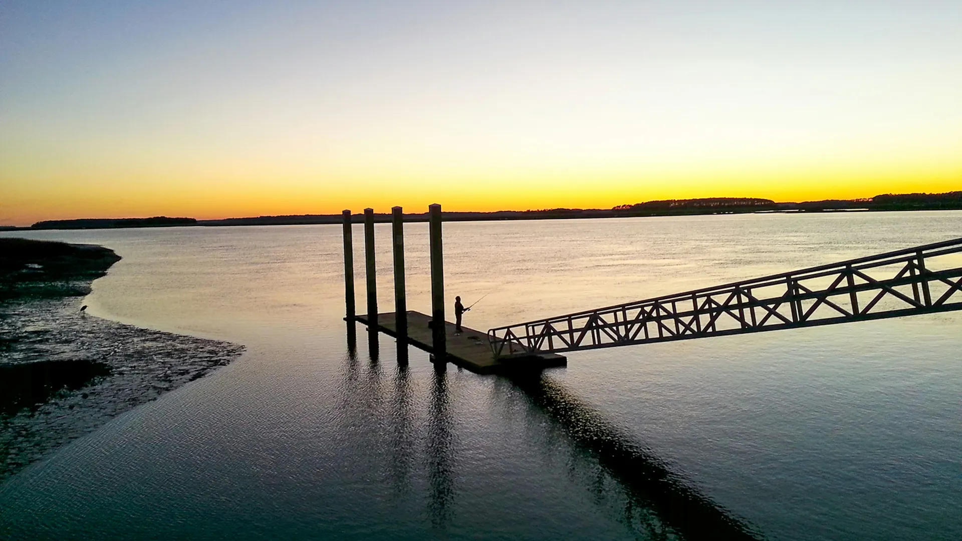 Silhouette of a person fishing on a dock at sunset with calm water and clear sky.