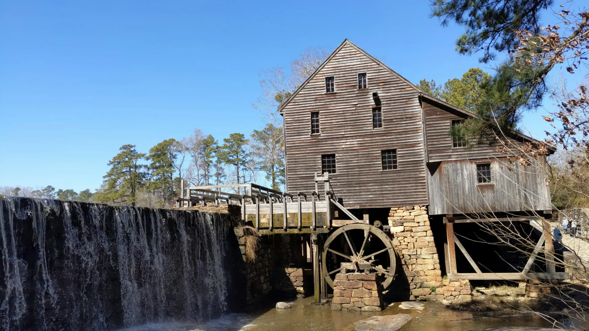 Historic wooden watermill with large wheel beside cascading waterfall under clear blue sky.