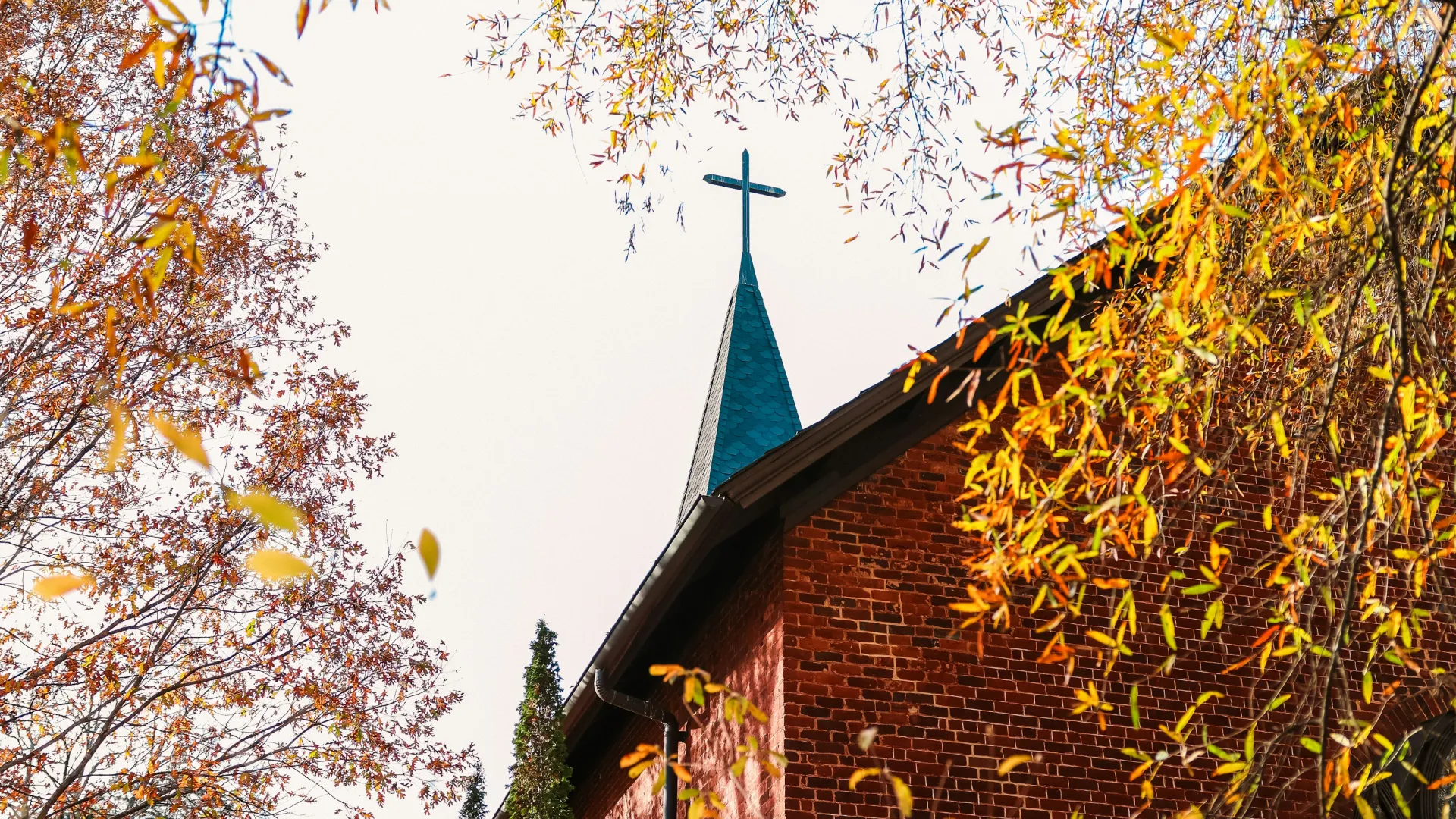 Autumn leaves frame a brick church with a cross-topped steeple against a pale sky.