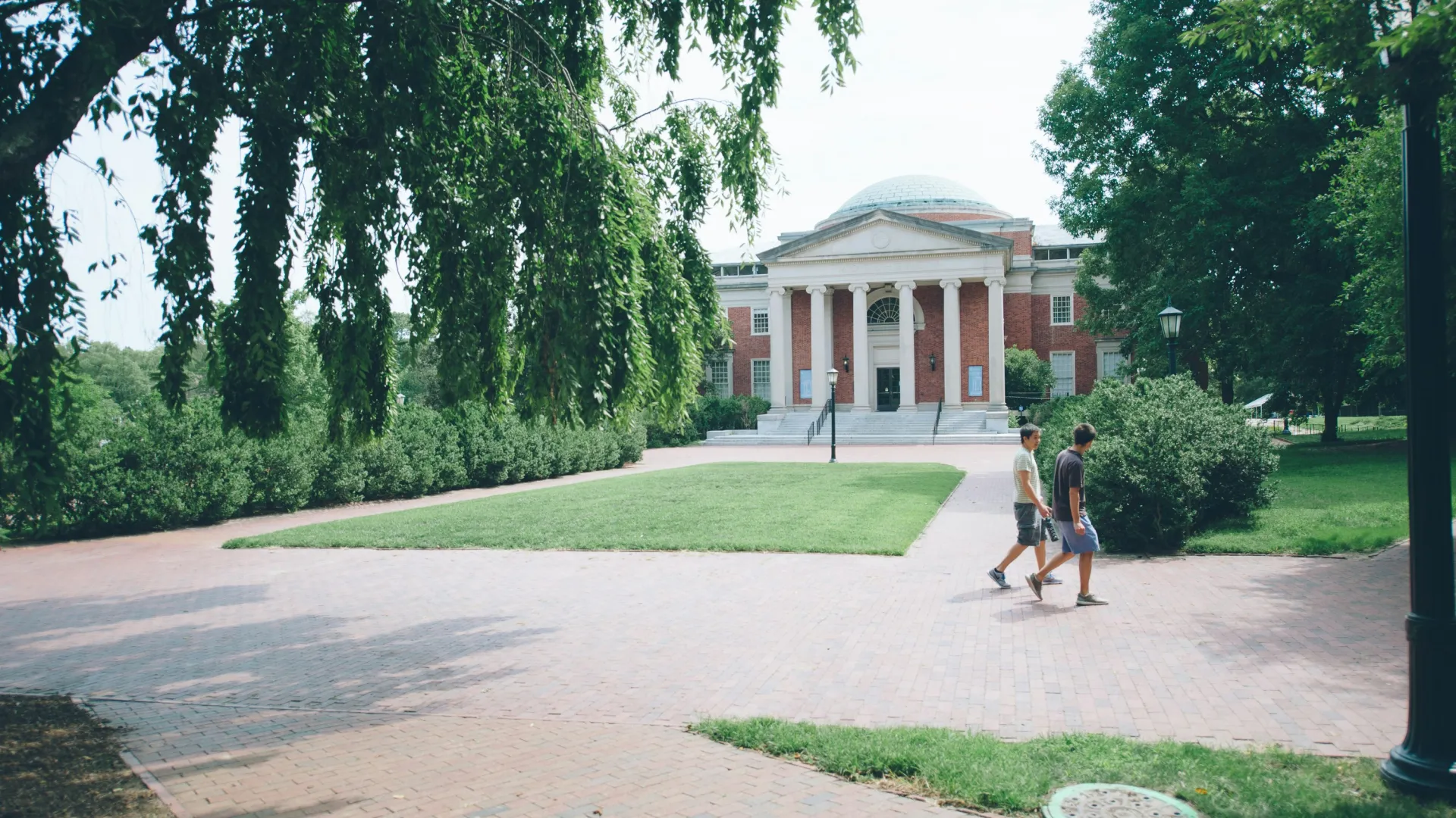 Two people walking on a brick path toward a classical university building with columns and dome surrounded by trees.