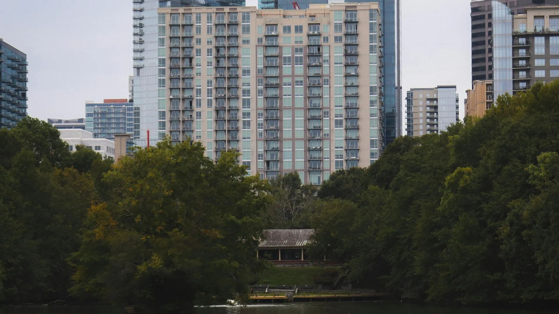 City skyline with modern buildings and apartments behind a river and dense green trees on an overcast day