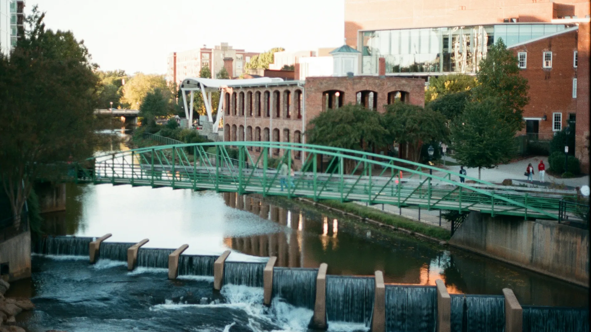 Green pedestrian bridge over flowing river with urban buildings and trees in the background on a sunny day