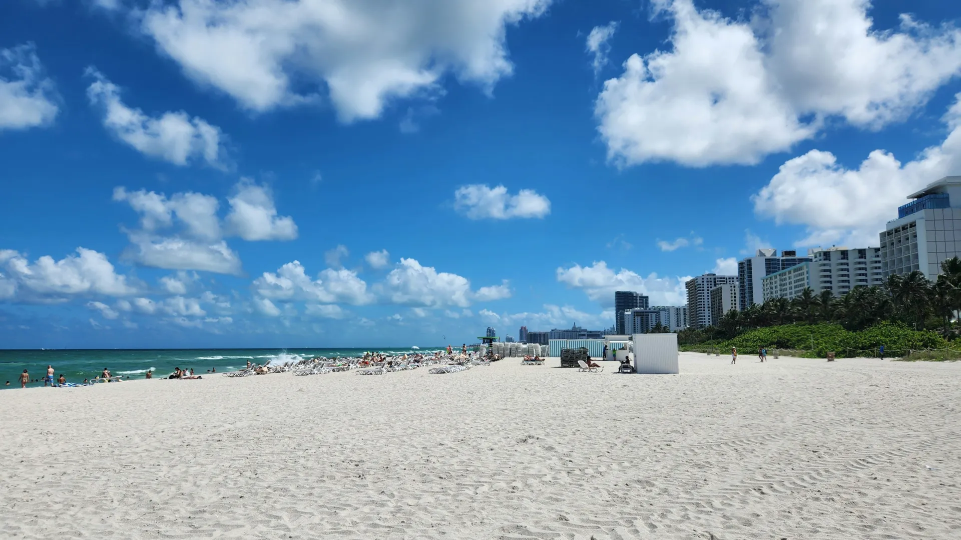 Sunny beach with white sand, blue ocean, scattered clouds, and beachfront hotels under clear sky.