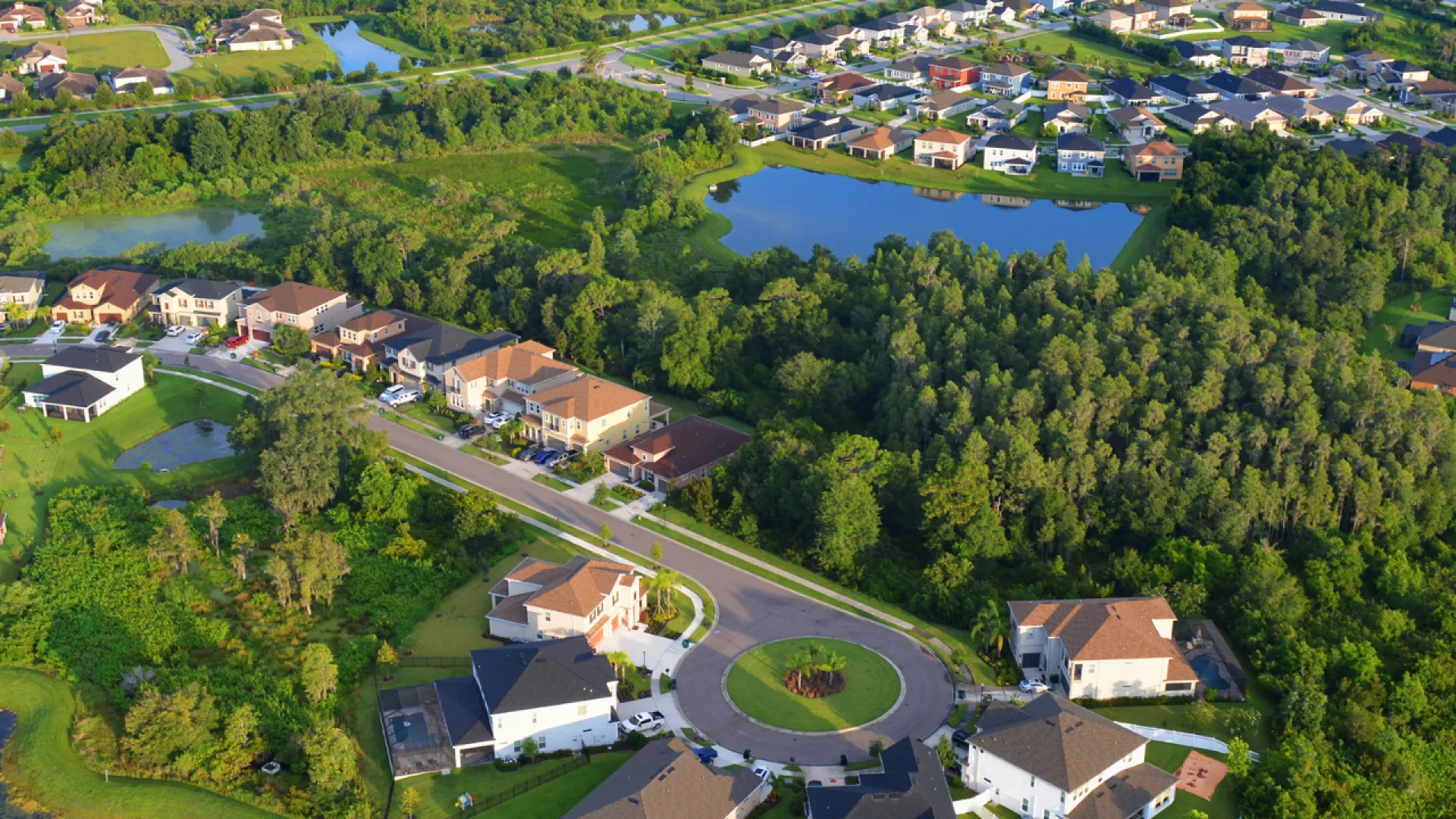 Aerial view of a suburban neighborhood with houses, green spaces, trees, and water bodies under daylight.
