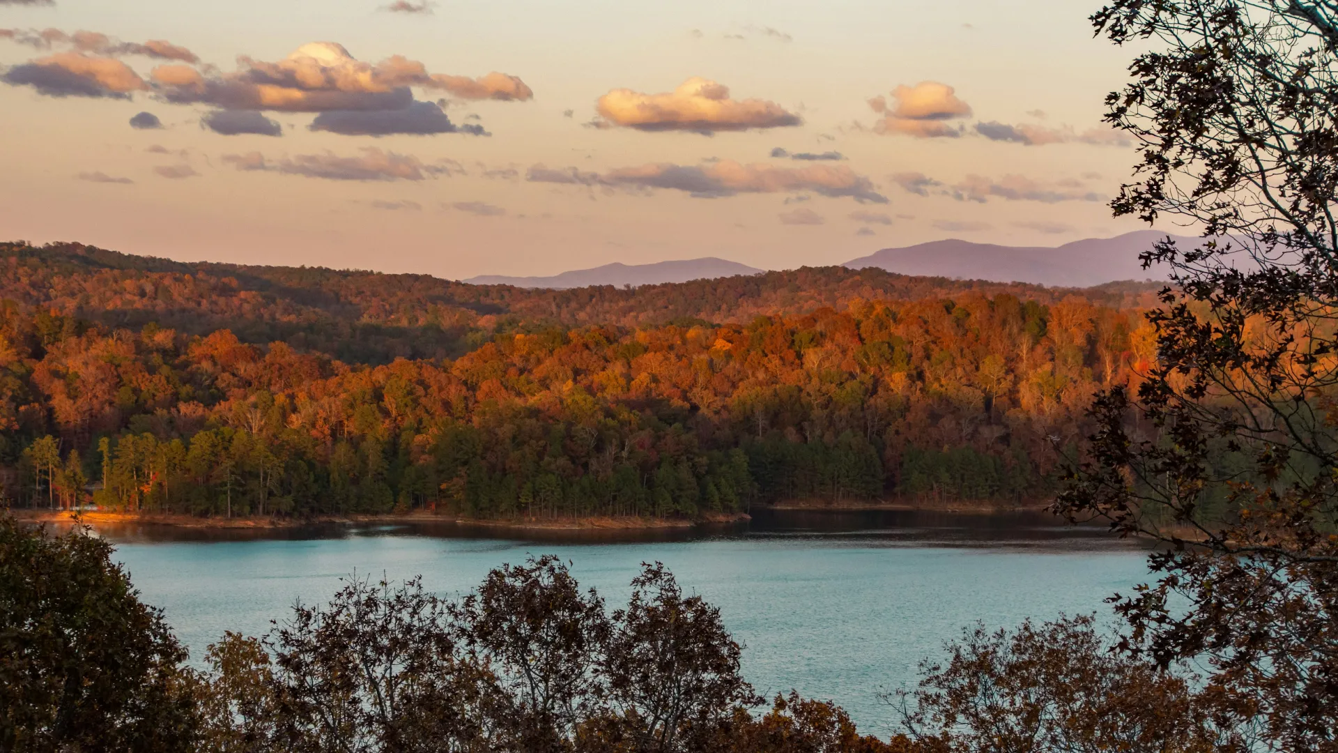 Scenic lake with autumn-colored trees on rolling hills under a partly cloudy sky during sunset.