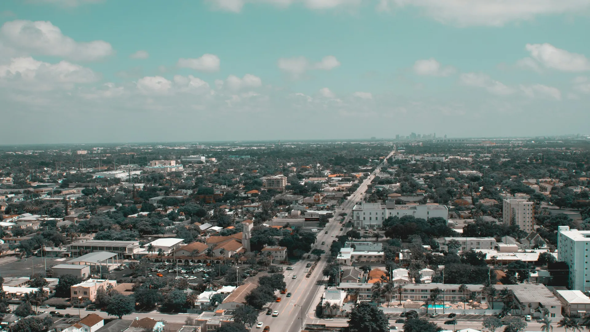 Aerial view of a city street stretching into the distance under a cloudy blue sky with surrounding buildings and vehicles.