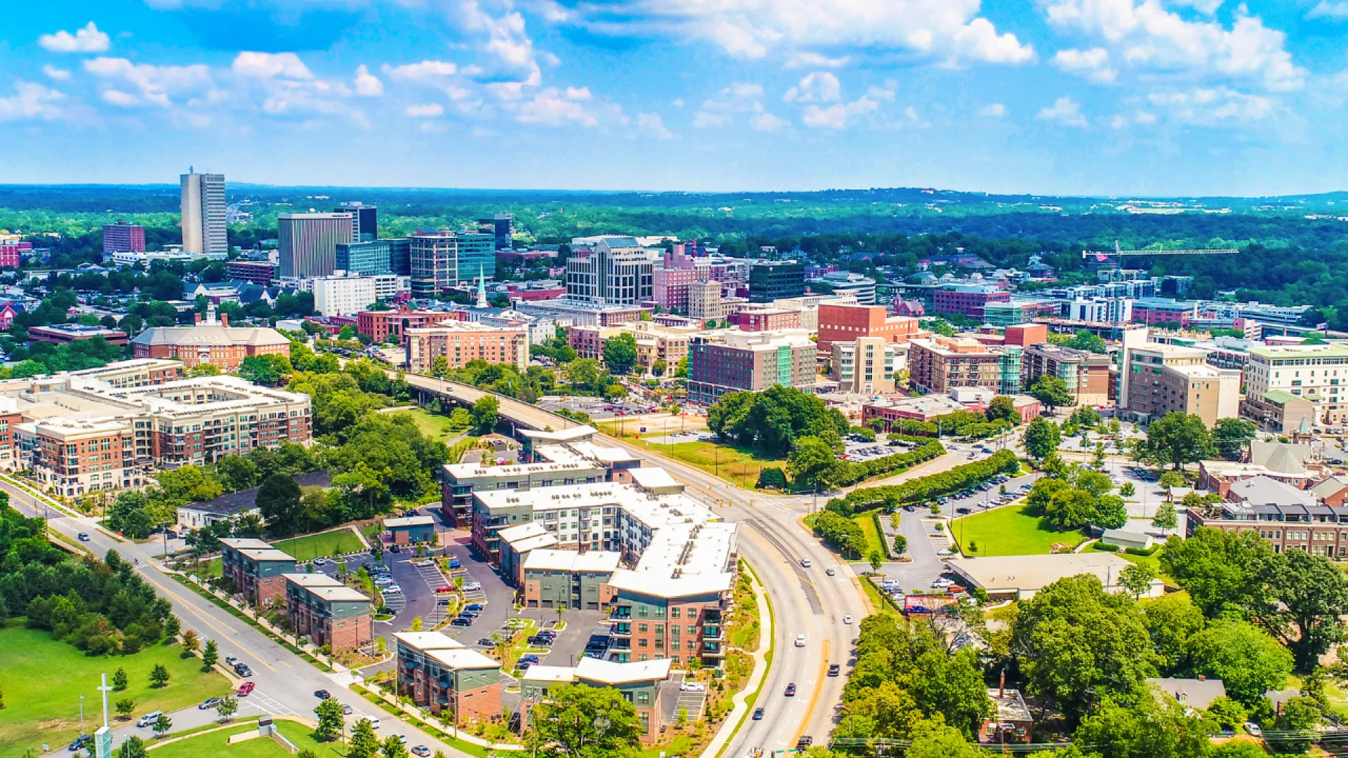 Aerial view of a modern cityscape with roads, buildings, and lush green trees under a clear sky.