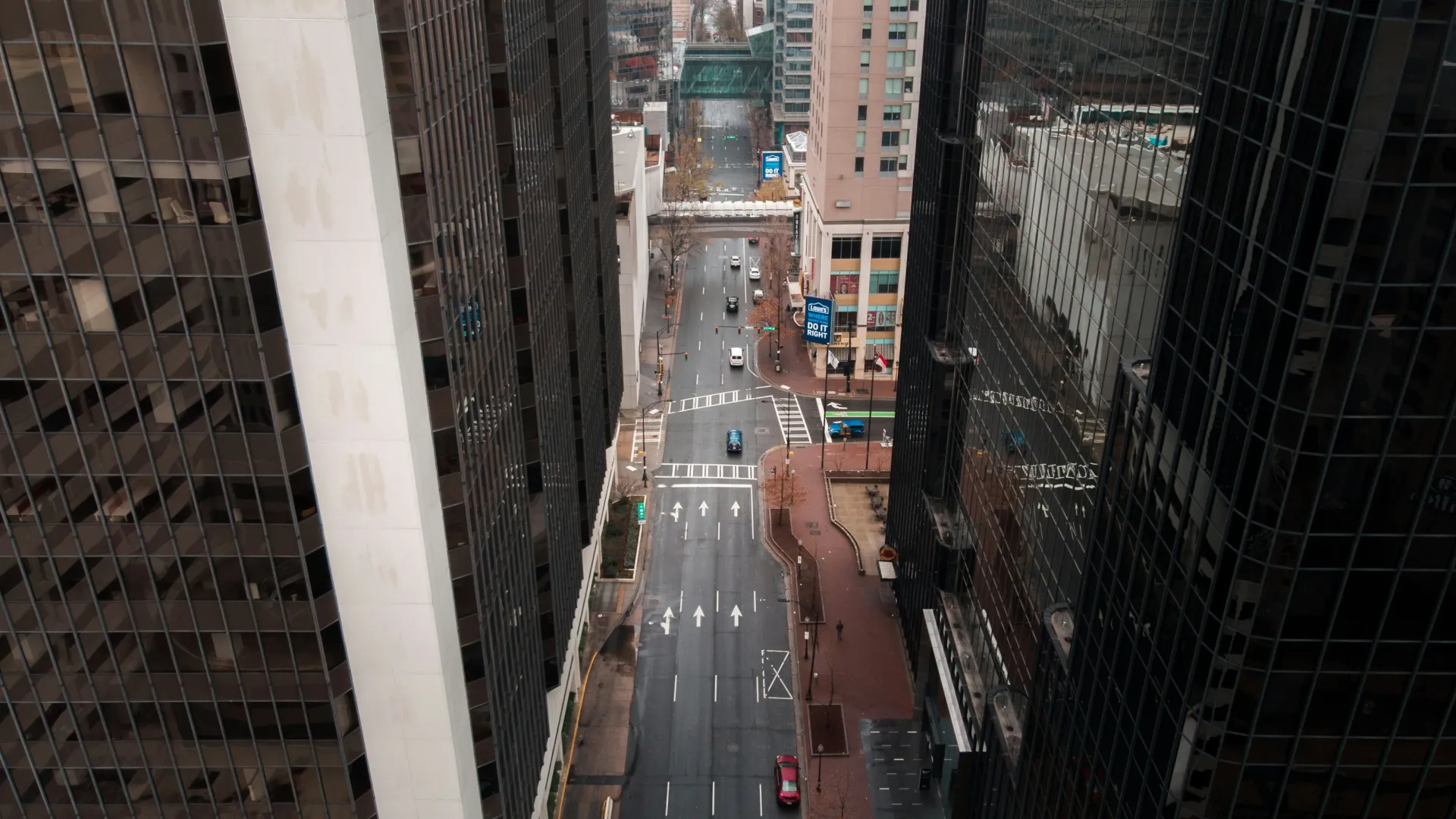 Aerial view of a city street flanked by tall glass skyscrapers with cars and pedestrians in an urban setting.