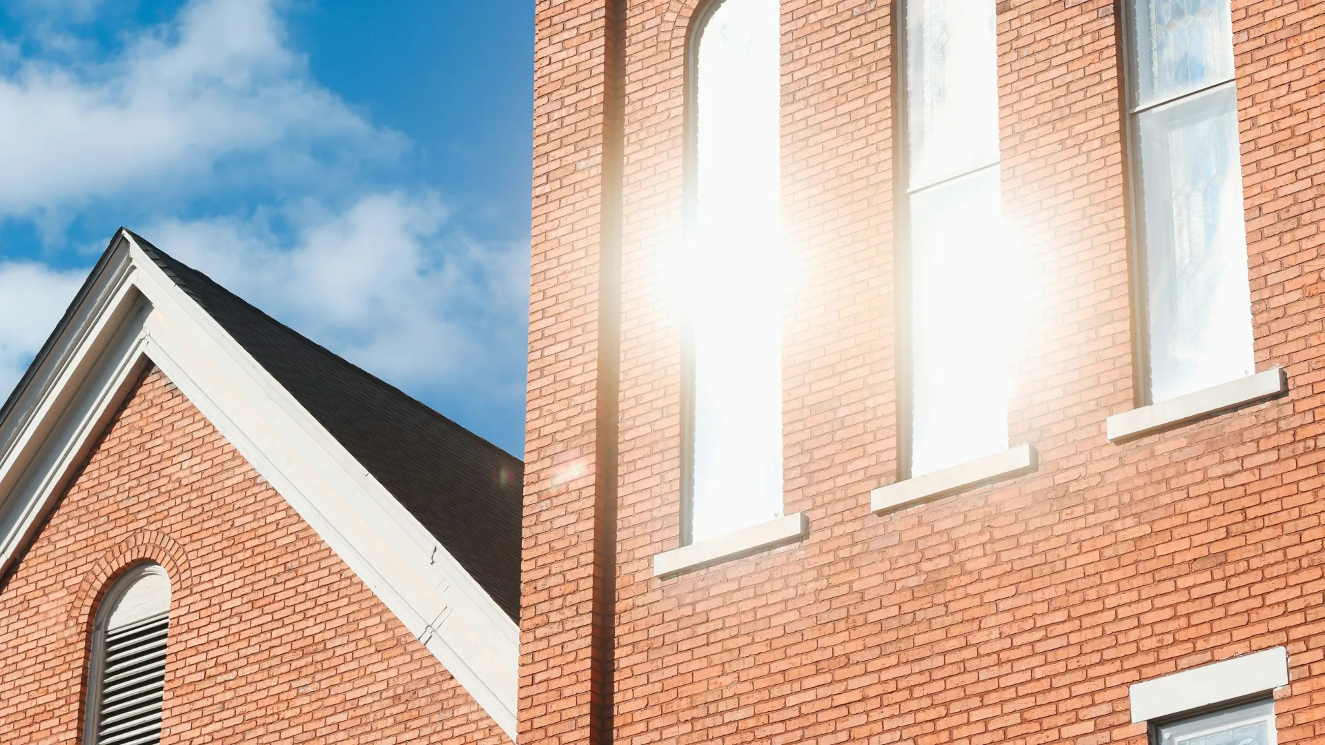 Sunlight reflects off tall arched windows of a red brick church building under a blue sky.