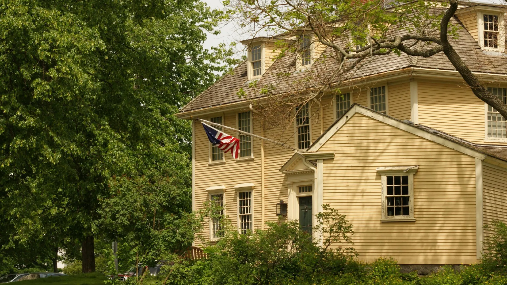 Historic yellow wooden building with American flag and large trees surrounding a peaceful garden area.
