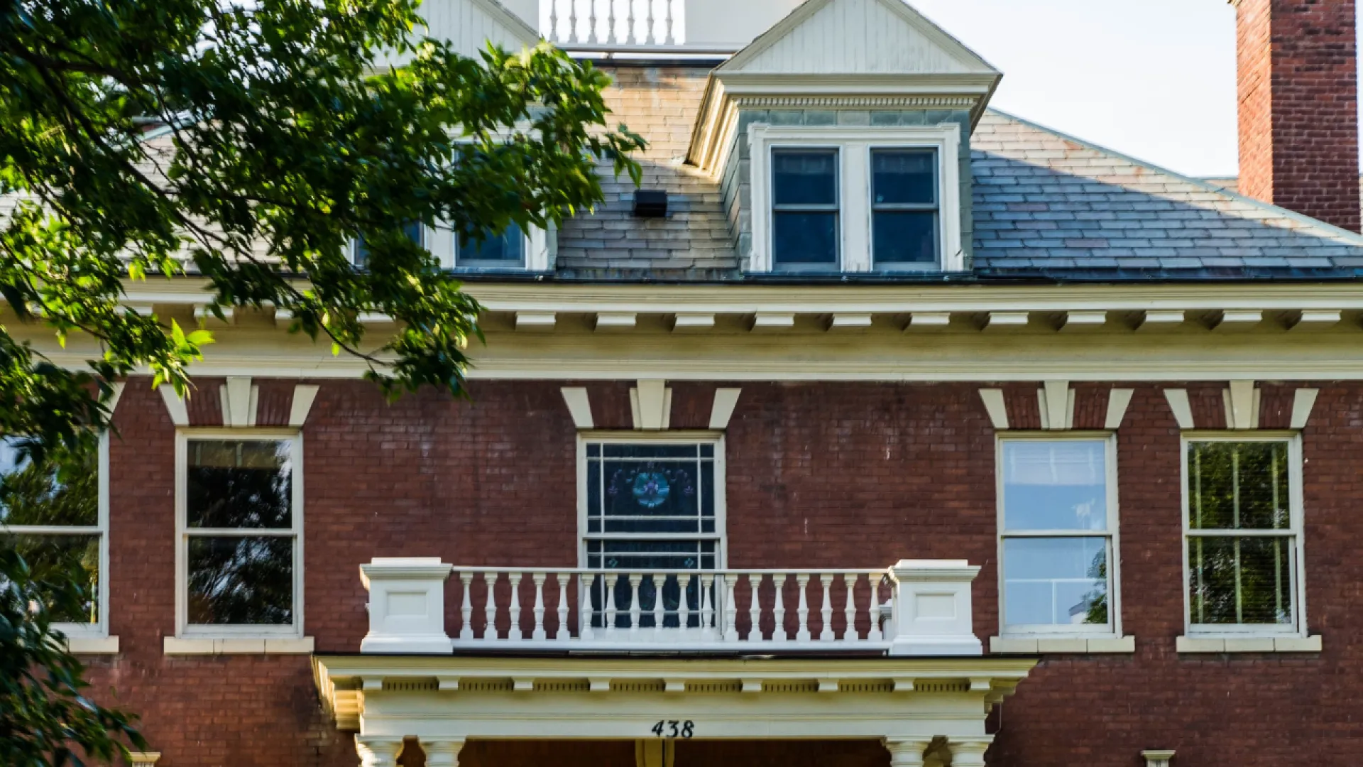 Historic brick mansion with white trim, dormer windows, and a stained glass window under leafy trees.