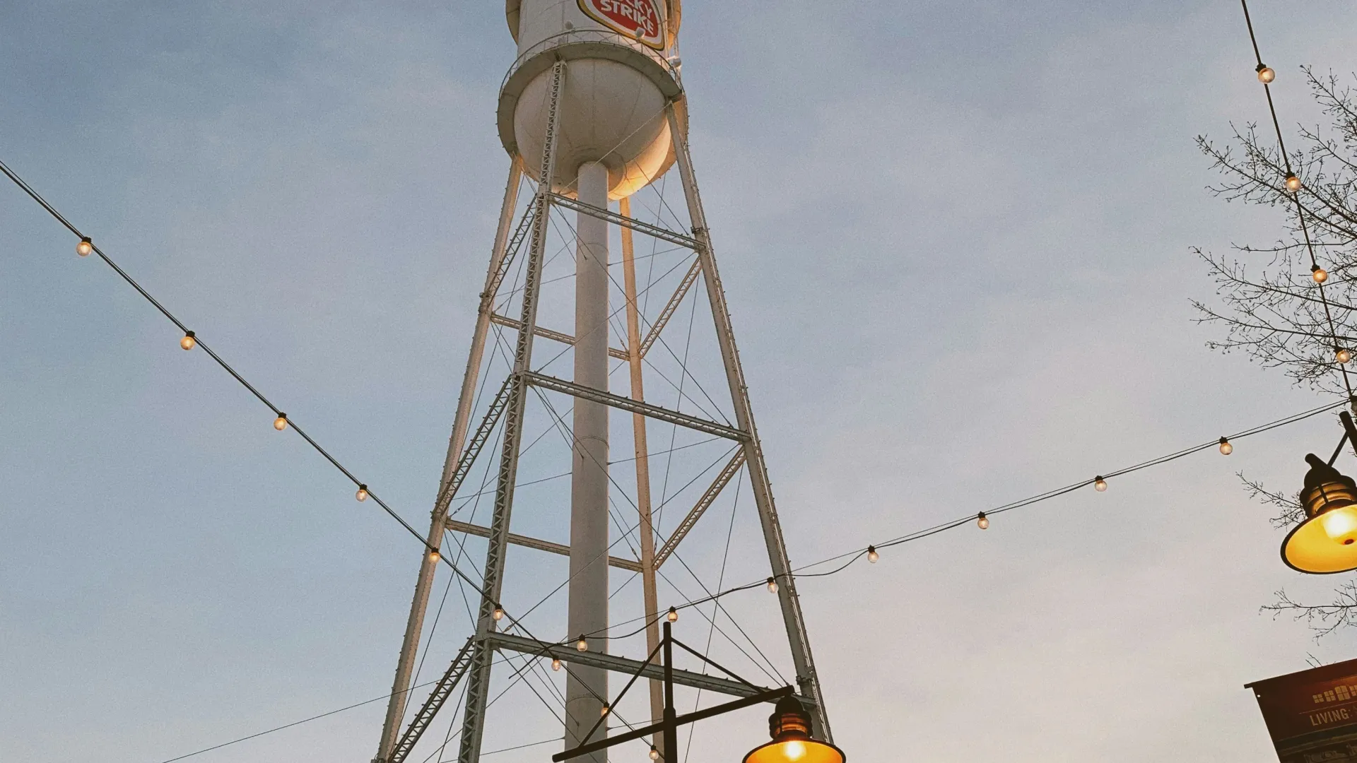 Tall Lucky Strike water tower at dusk with string lights and street lamps glowing under a clear sky.