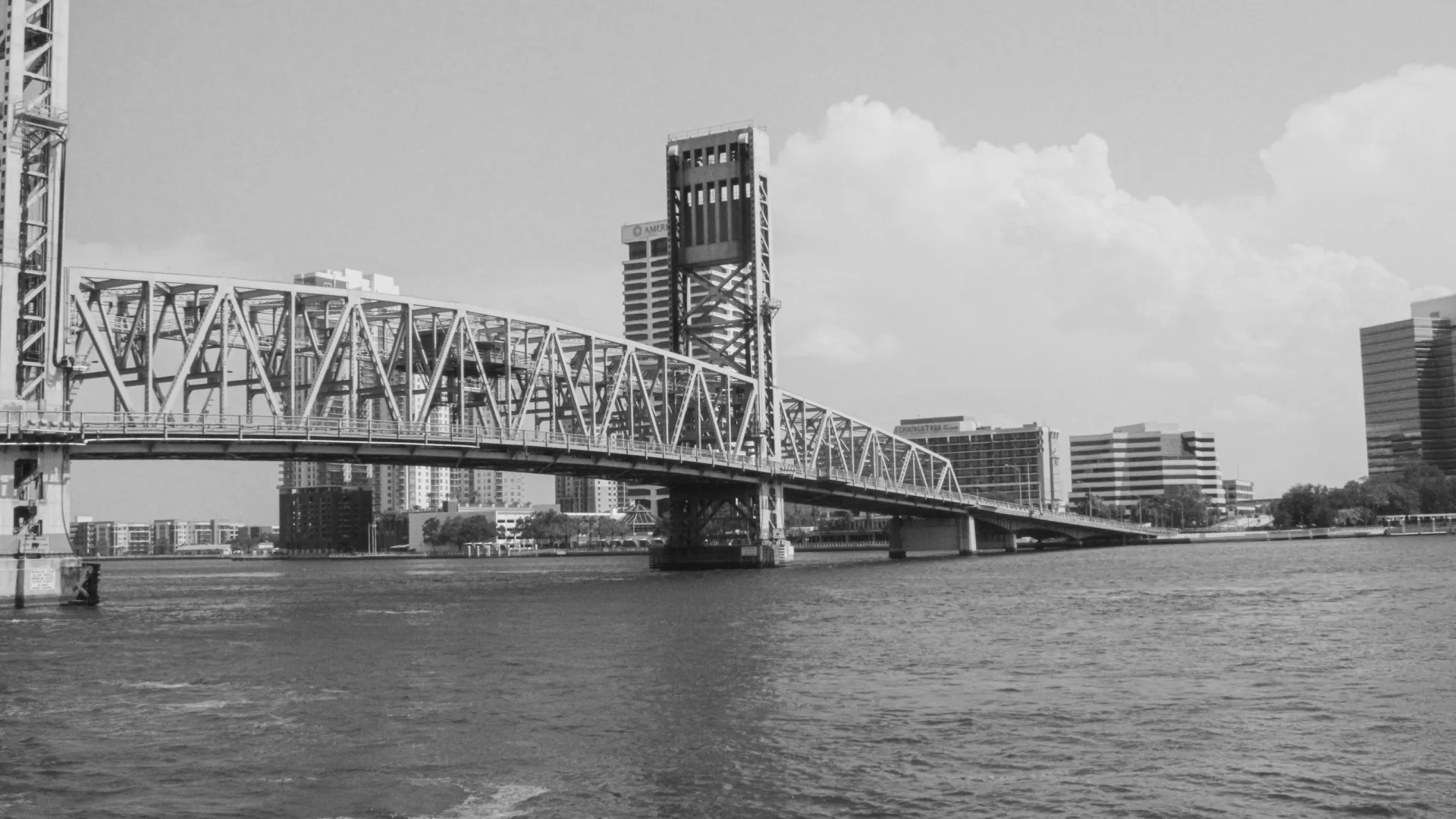 Black and white image of a metal truss drawbridge over a river with city buildings in the background on a cloudy day