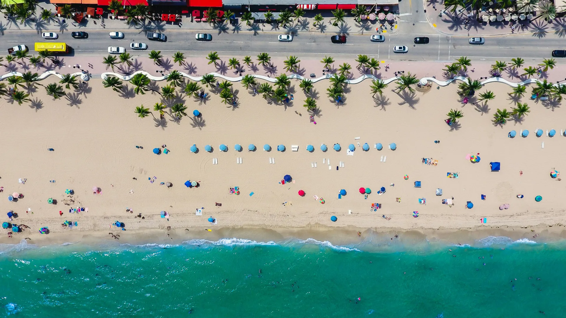 Aerial view of a sunny beach with umbrellas, palm trees, people, and clear turquoise water along the shore.