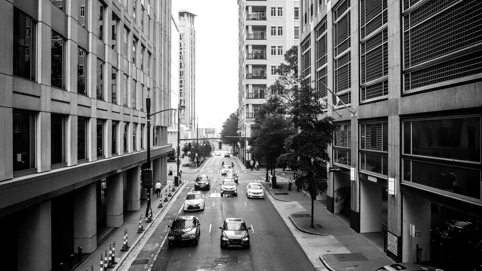 Black and white city street scene with cars driving between tall office buildings on an urban road.