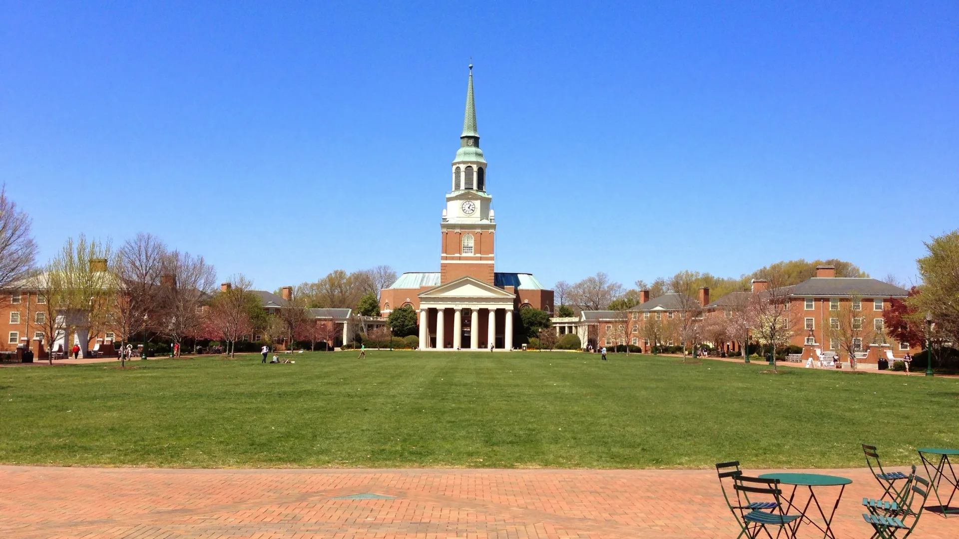 Green lawn and brick plaza with tables in front of a central building with clock tower and steeple under clear blue sky