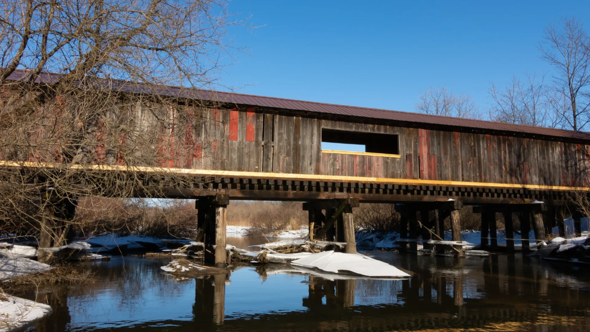 Rustic wooden covered bridge over a calm river with snow and leafless trees under clear blue sky.