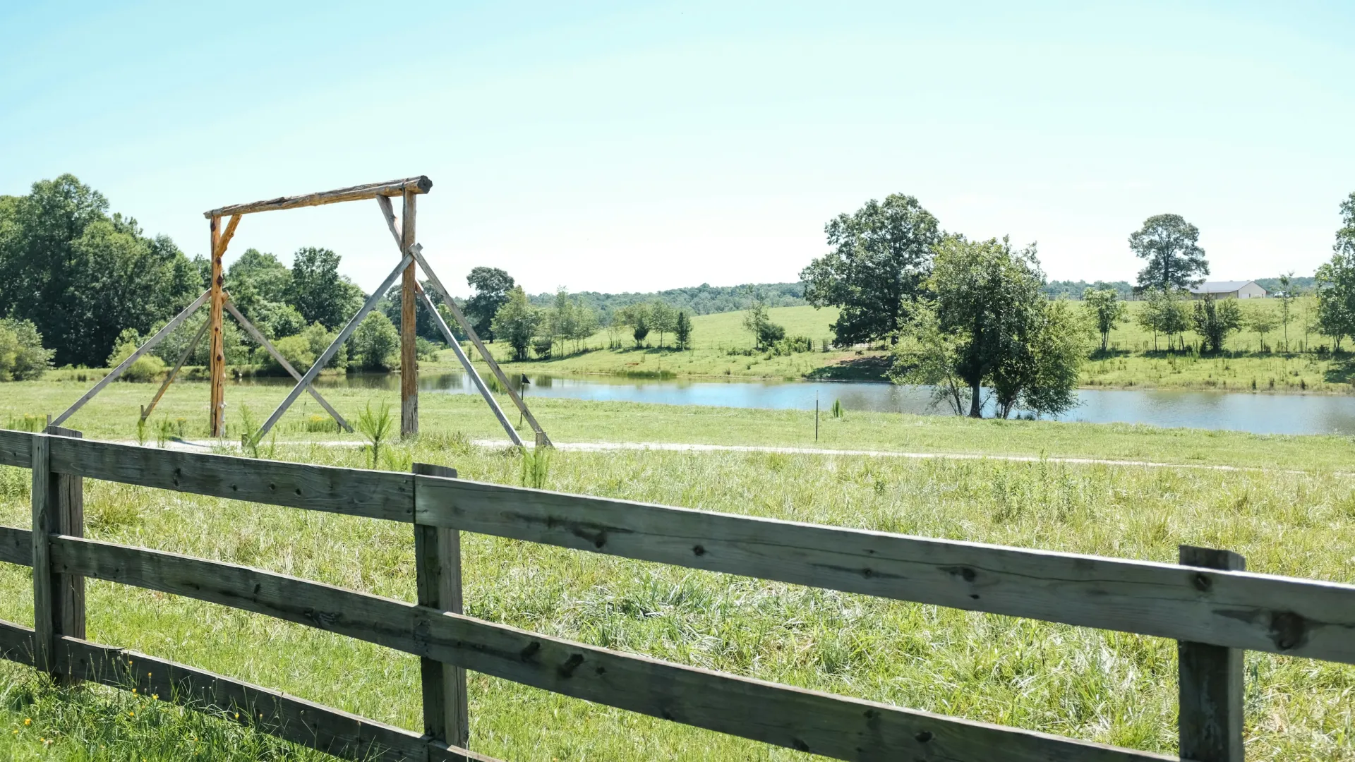 Wooden fence on a green grassy field with a pond and trees under a clear blue sky.