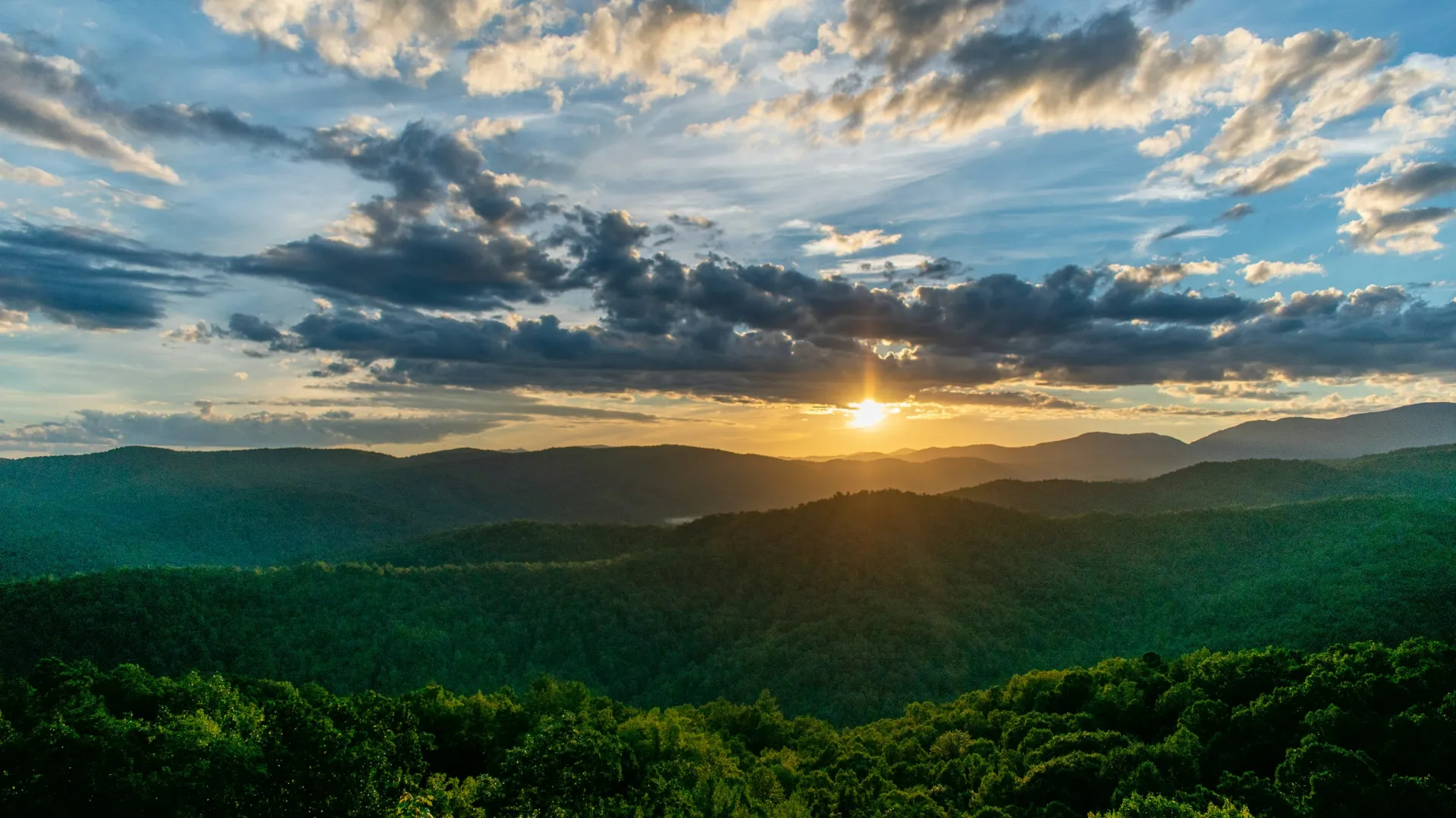 Sunset over green forested mountains under a sky filled with dramatic clouds and sunlight beams.