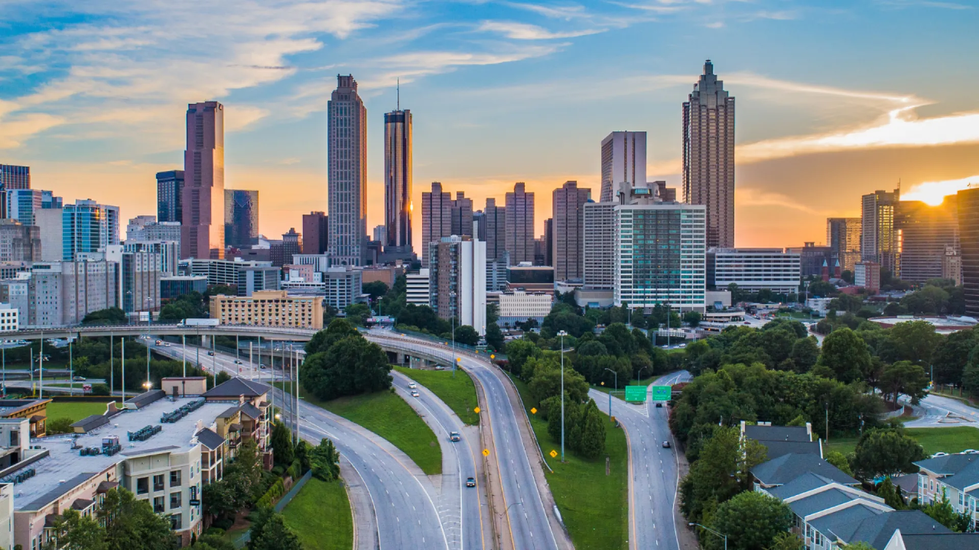 Sunset over a modern city skyline with tall buildings, highways, and green trees in the foreground.