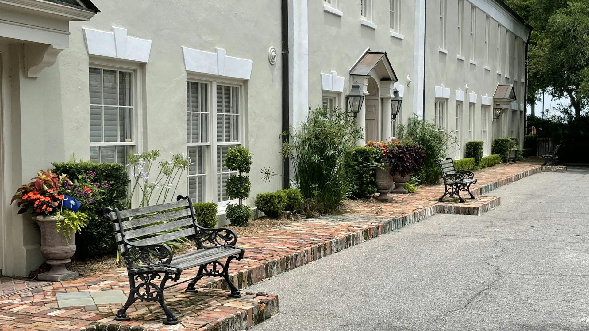 Black wrought iron benches on brick sidewalk beside beige building with decorative plants and windows.