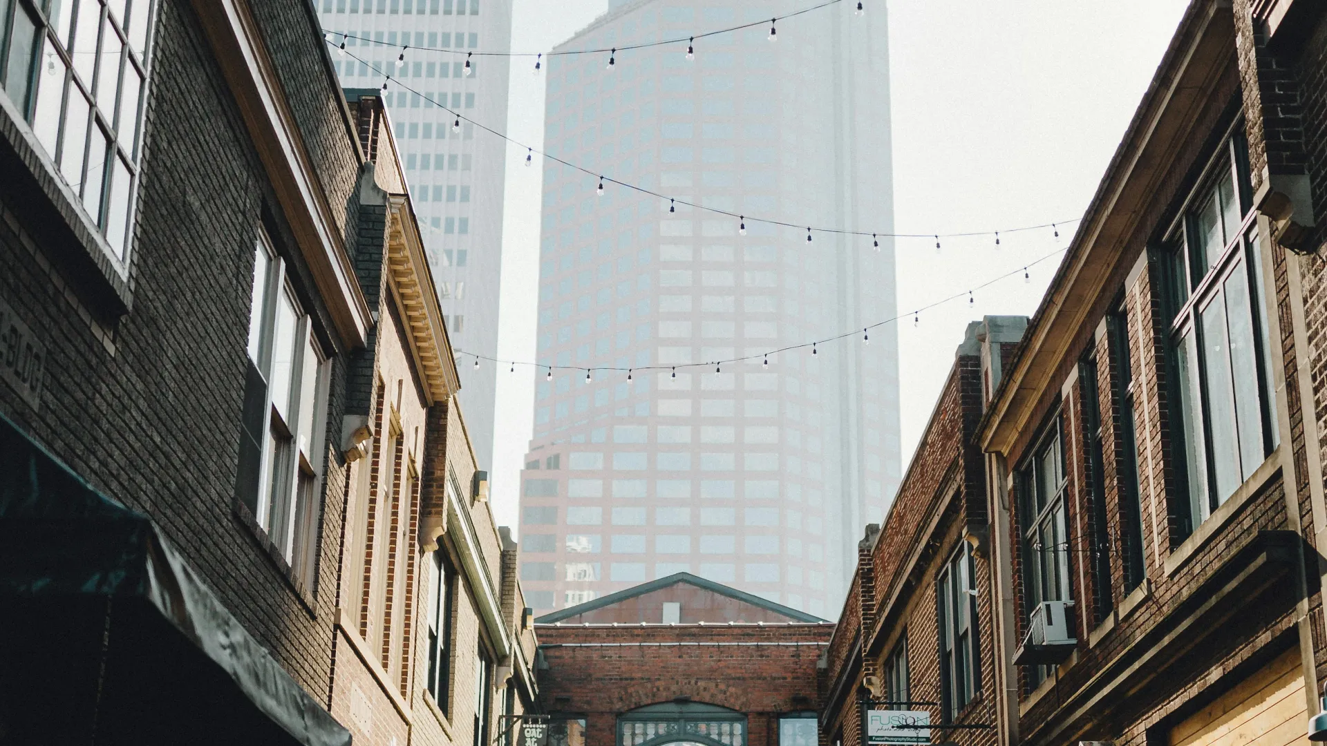 Sunny urban alley with brick buildings, string lights overhead, and a distant modern skyscraper on a clear day.