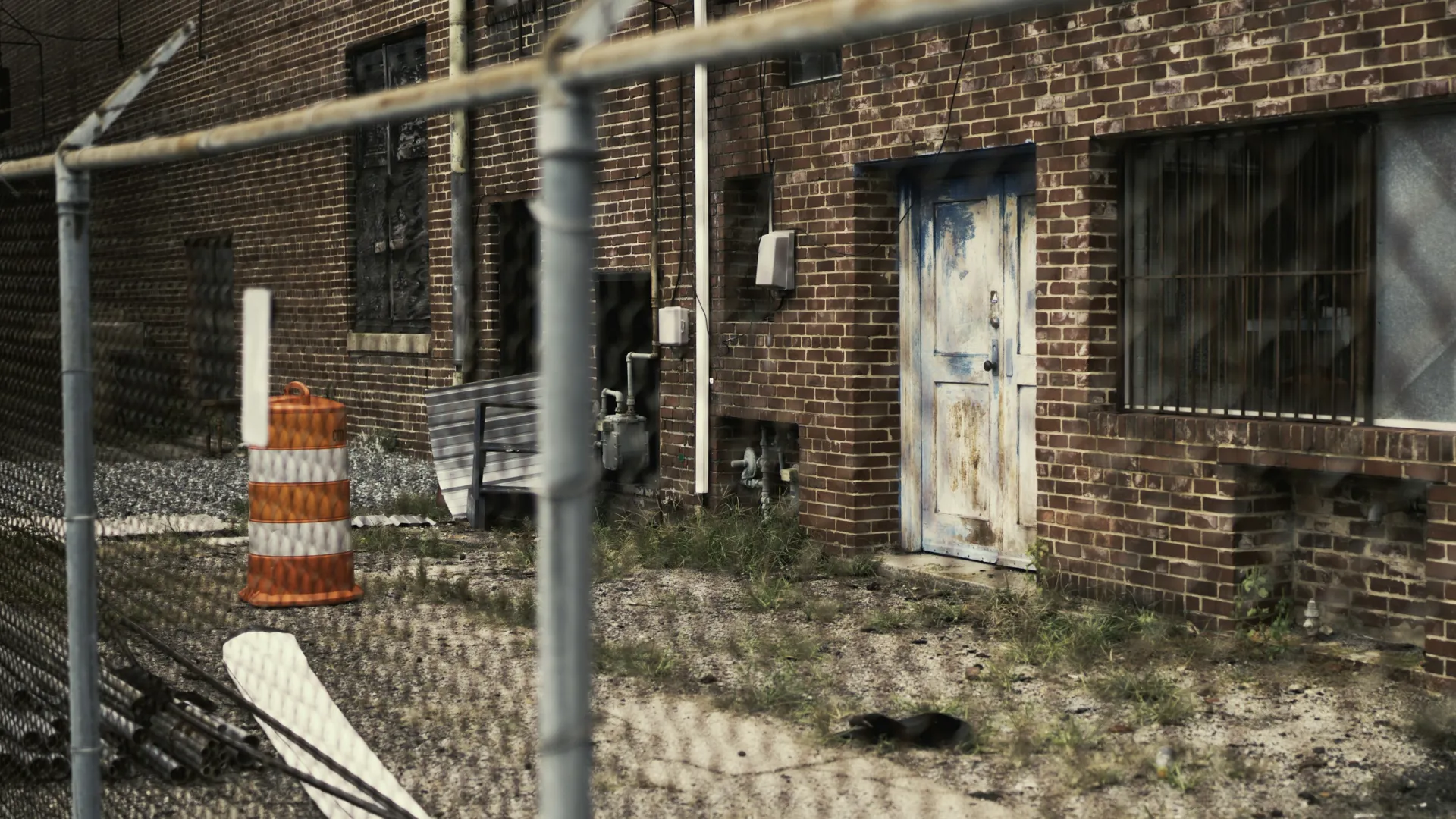 Rusty white door and barred windows on brick building behind chain-link fence with construction barrel