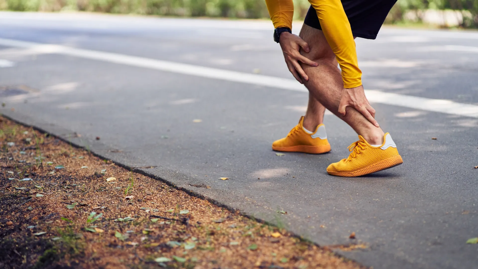 Runner wearing bright orange shoes and yellow shirt holding ankle in pain on outdoor path.