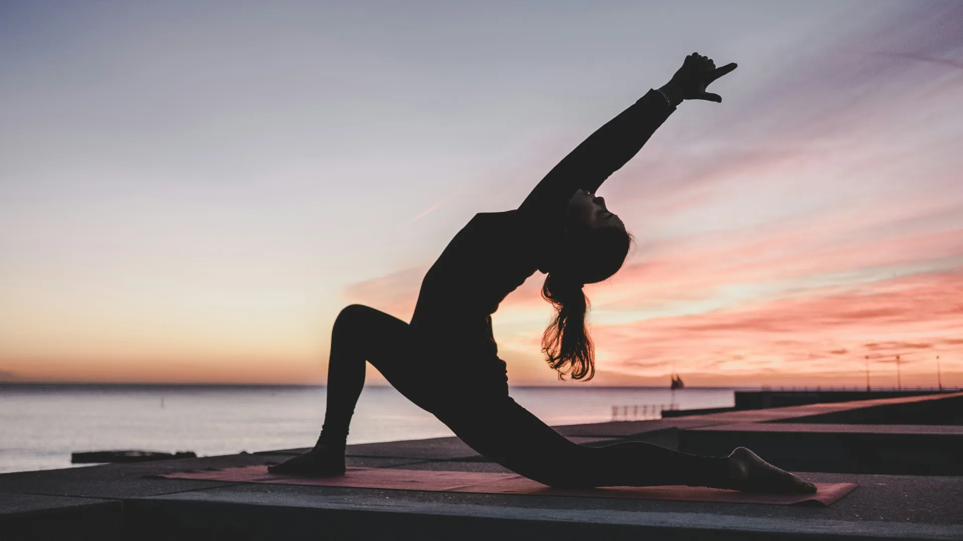 Woman practicing yoga on a mat by the sea during a colorful sunset with a peaceful sky.