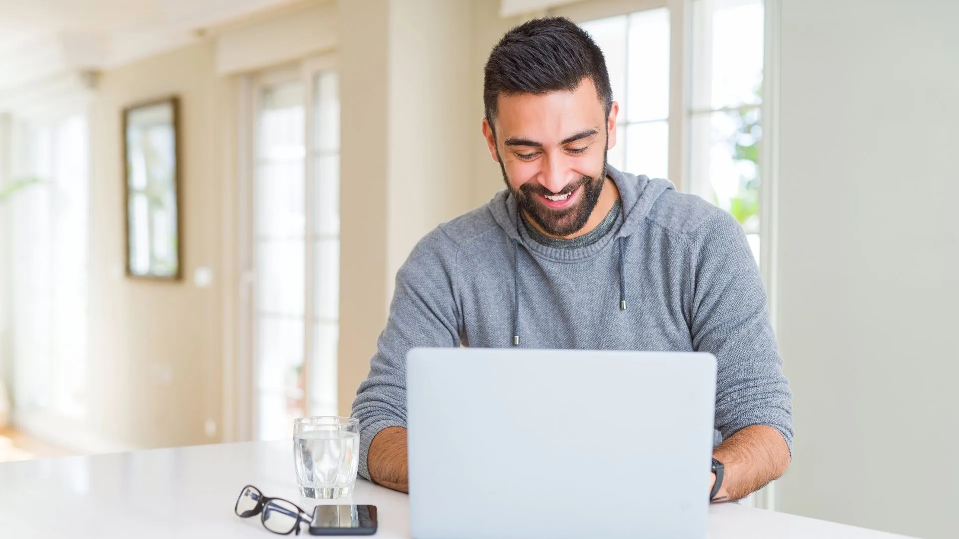 Smiling man using laptop at home with smartphone, glasses, and glass of water on white table