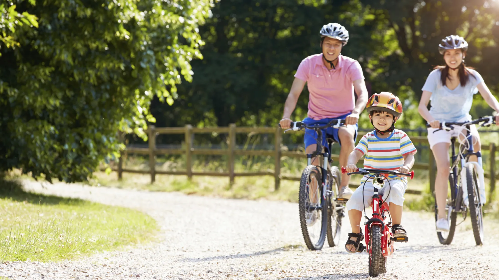 Family of three wearing helmets riding bikes on a sunny dirt path surrounded by trees and wooden fence.