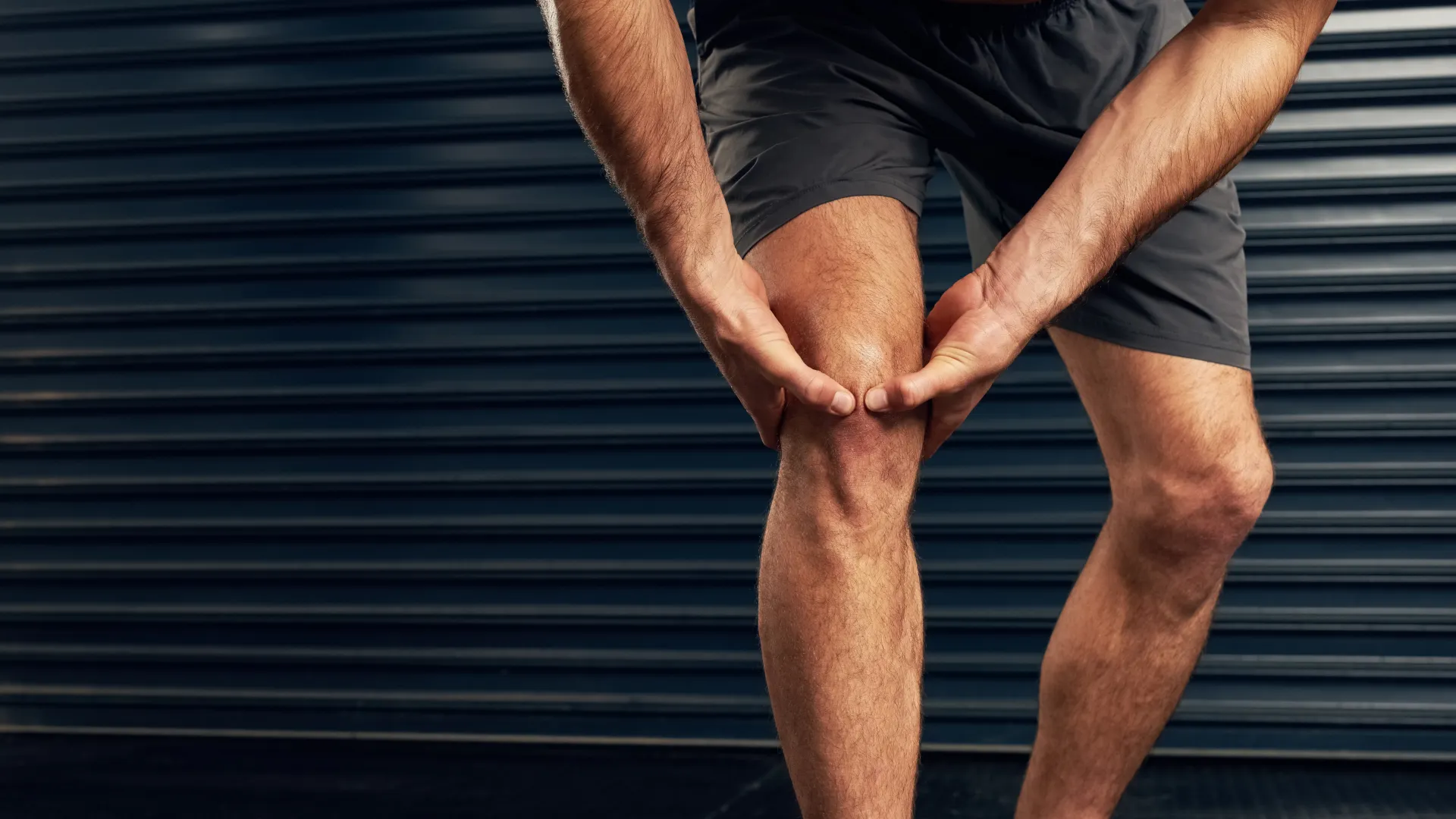 Athlete wearing black shorts holding knee in pain against dark industrial background