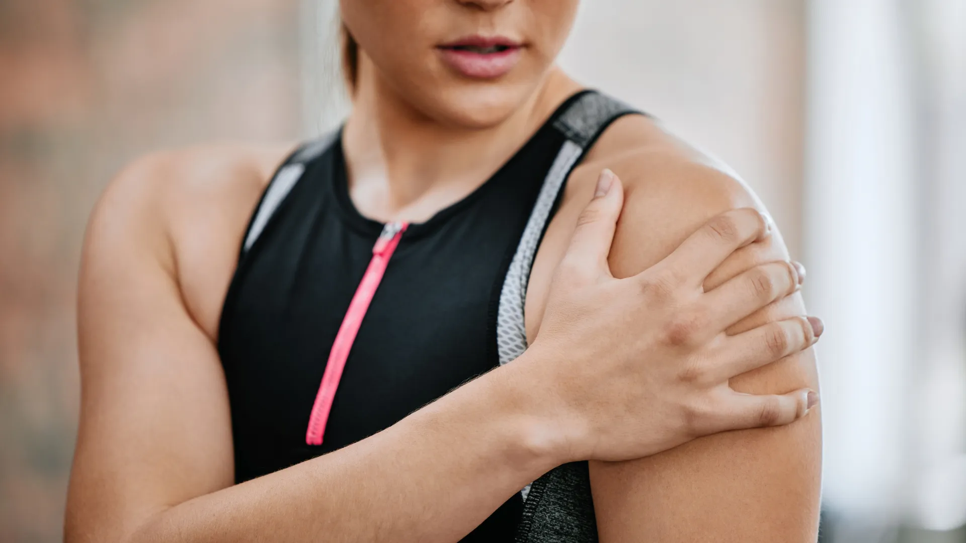 Close-up of a woman in workout gear holding her shoulder, indicating shoulder pain or injury post-exercise.