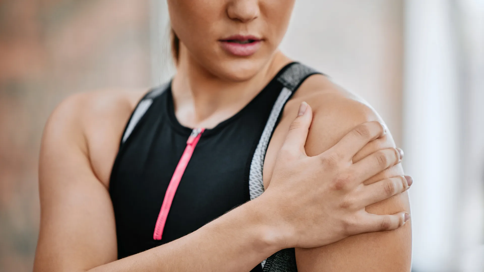 Close-up of a woman in workout gear holding her shoulder, indicating shoulder pain or injury post-exercise.