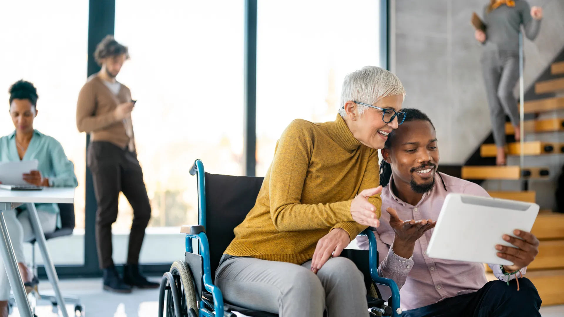 Smiling diverse coworkers collaborate with a tablet around a woman in wheelchair in a modern office space.