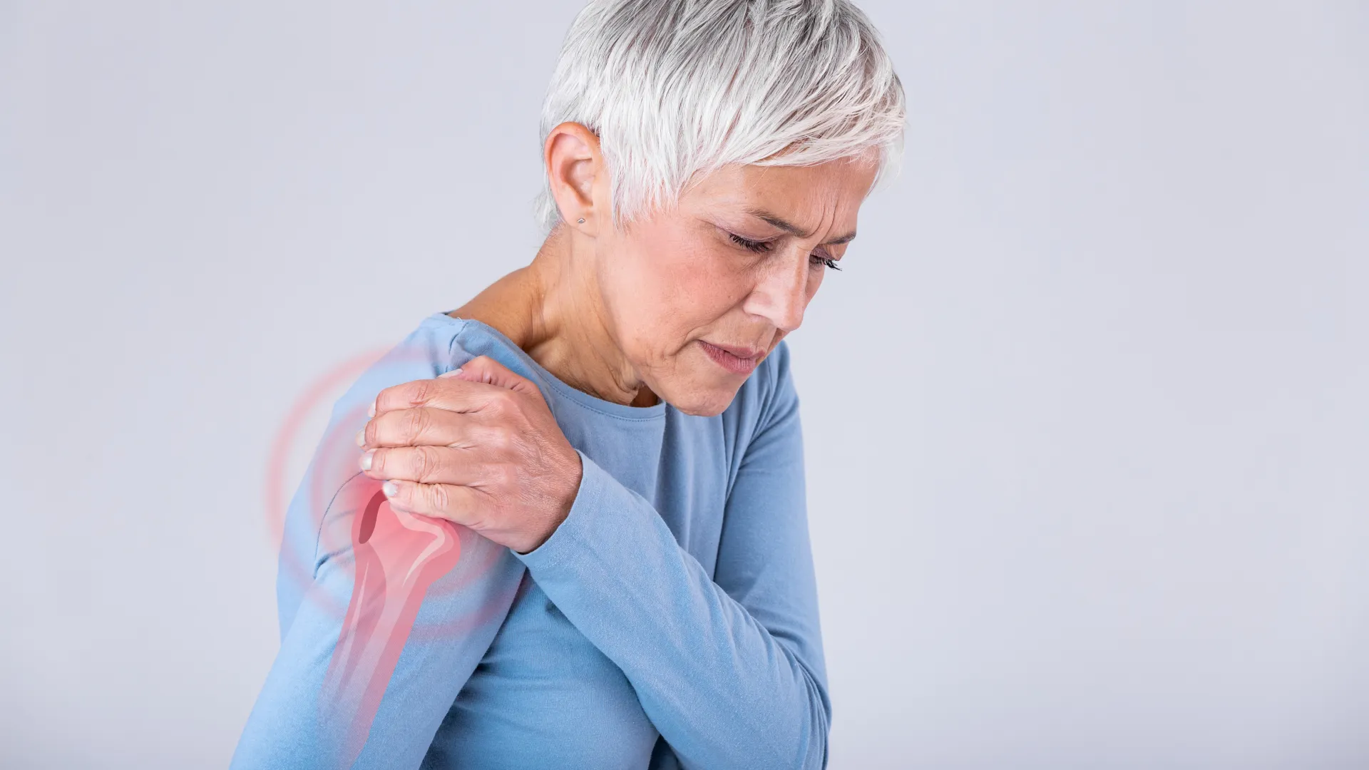 Middle-aged woman in blue shirt holding her shoulder in pain with red area highlighting discomfort.
