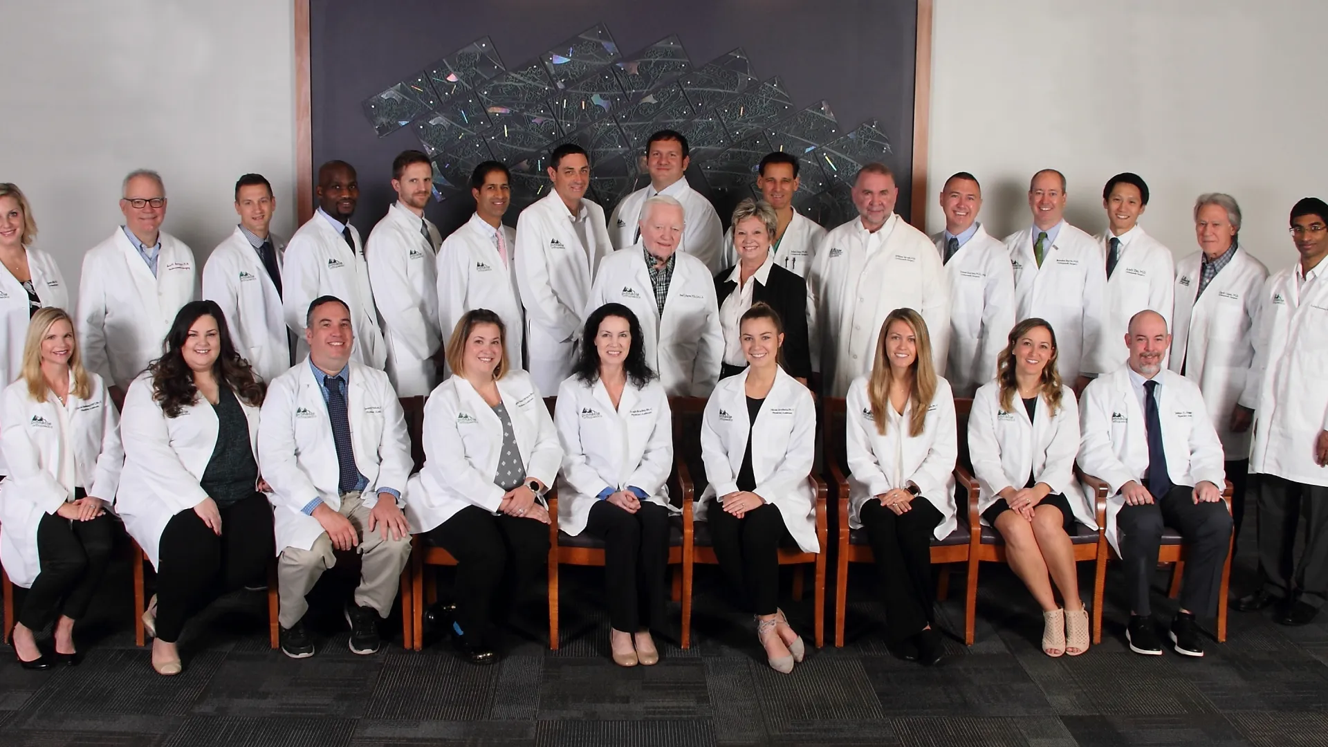 Group portrait of diverse medical professionals wearing white coats in a formal indoor setting.