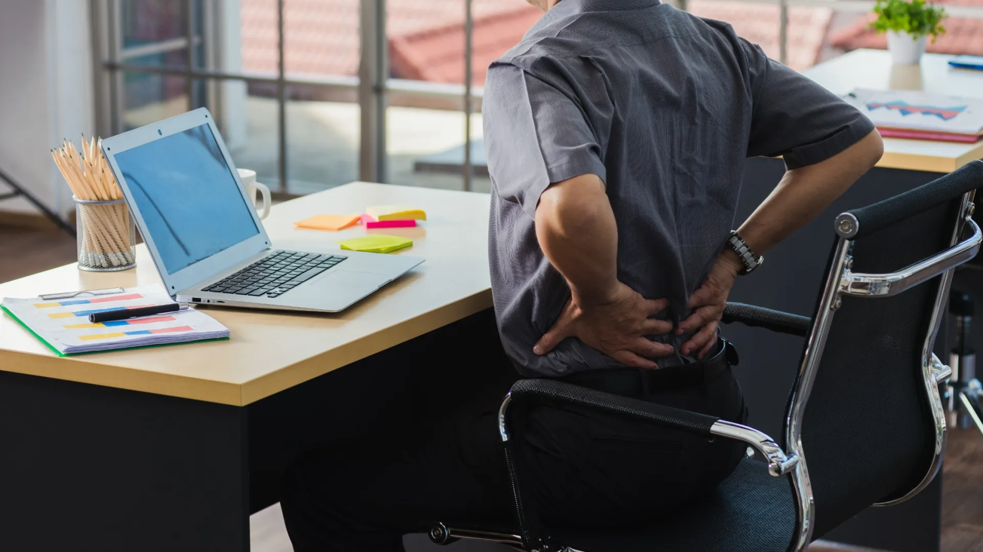 Man in office chair holding lower back, showing discomfort from back pain while working at desk with laptop.