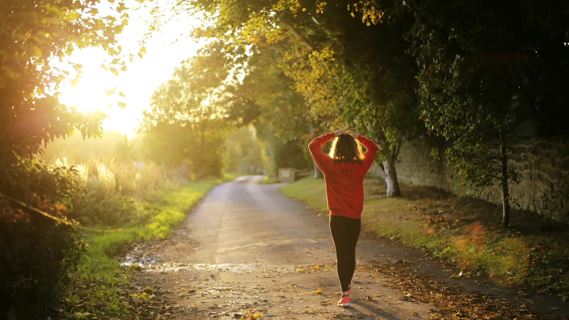 Woman in red jacket walking on a sunlit country road surrounded by trees and autumn leaves.