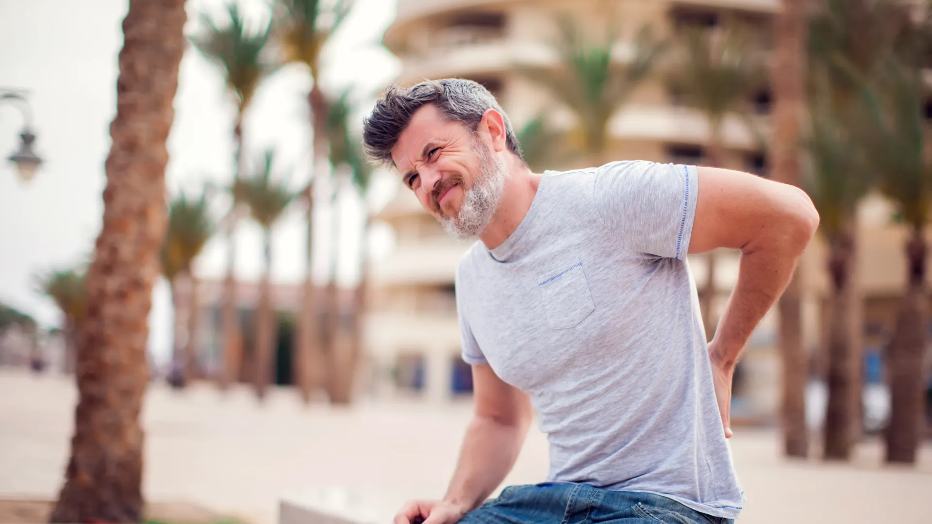 Middle-aged man in casual clothes sitting outdoors holding his lower back in pain near palm trees.