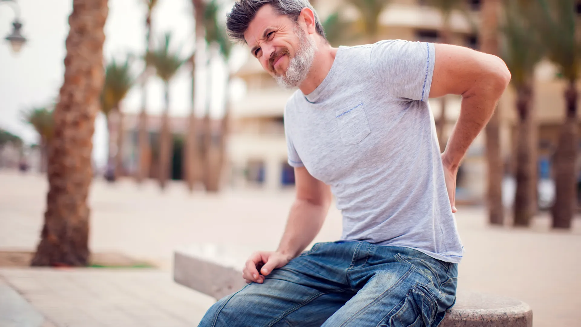 Middle-aged man in casual clothes sitting outdoors holding his lower back in pain near palm trees.