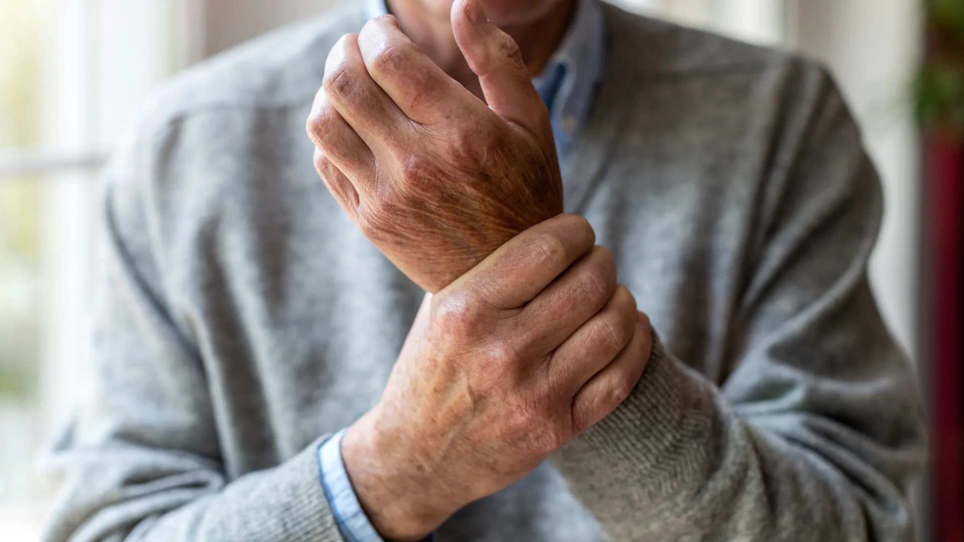 Elderly man in gray sweater holding his wrist, indicating wrist pain or discomfort indoors near a window.