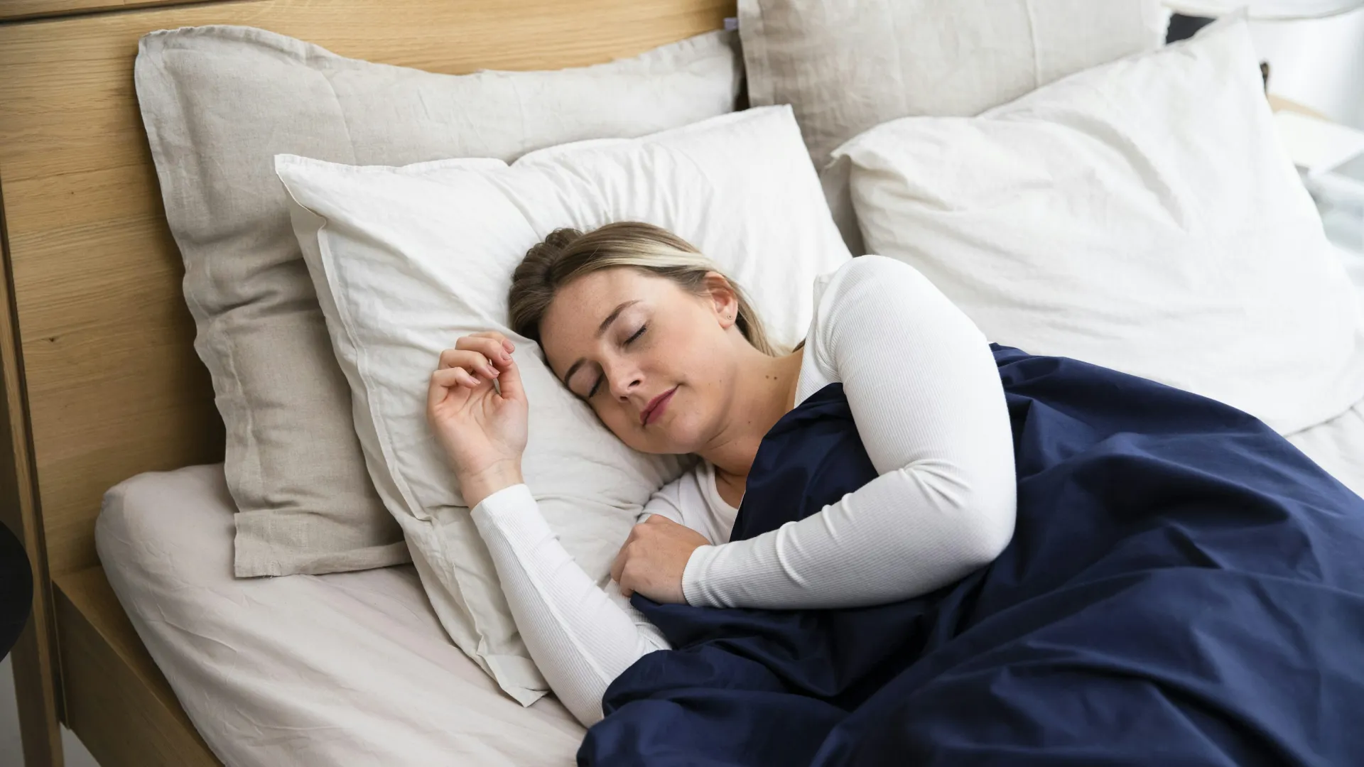 Woman sleeping peacefully in bed with white sheets and navy blue blanket in a wooden frame bed.