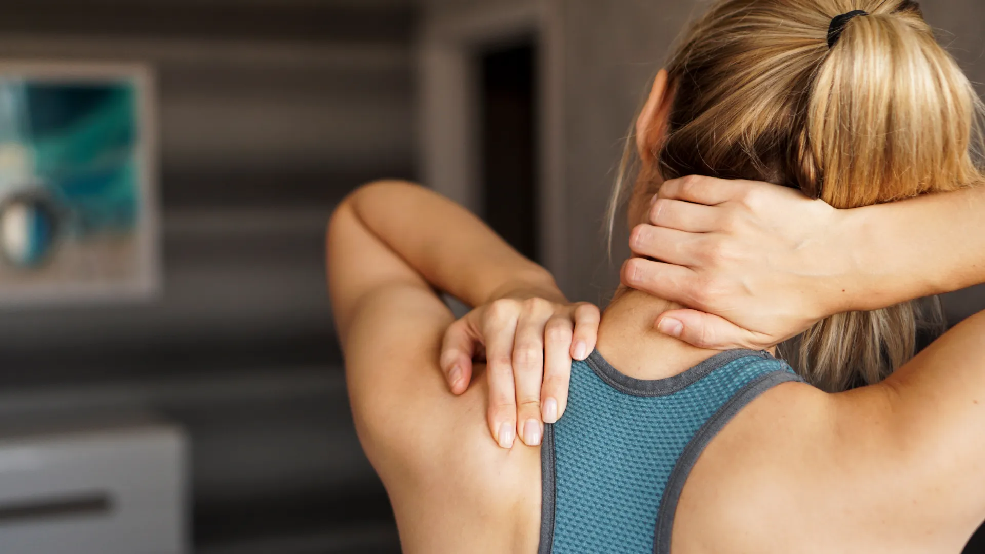 Woman in sportswear massaging her neck and upper back indoors, showing muscle tension relief.