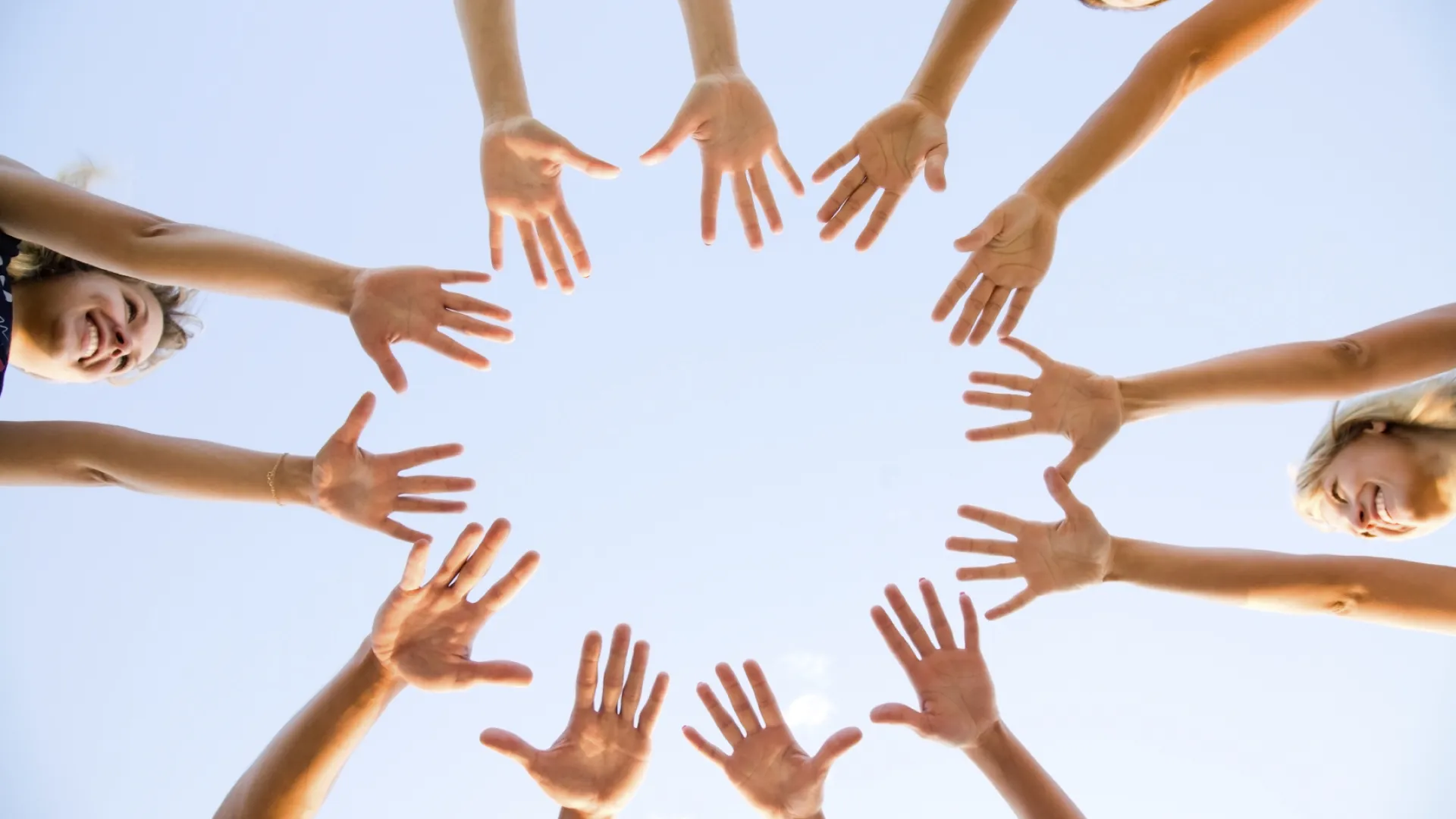 Group of diverse people reaching hands together in a circle under clear blue sky, symbolizing unity and teamwork