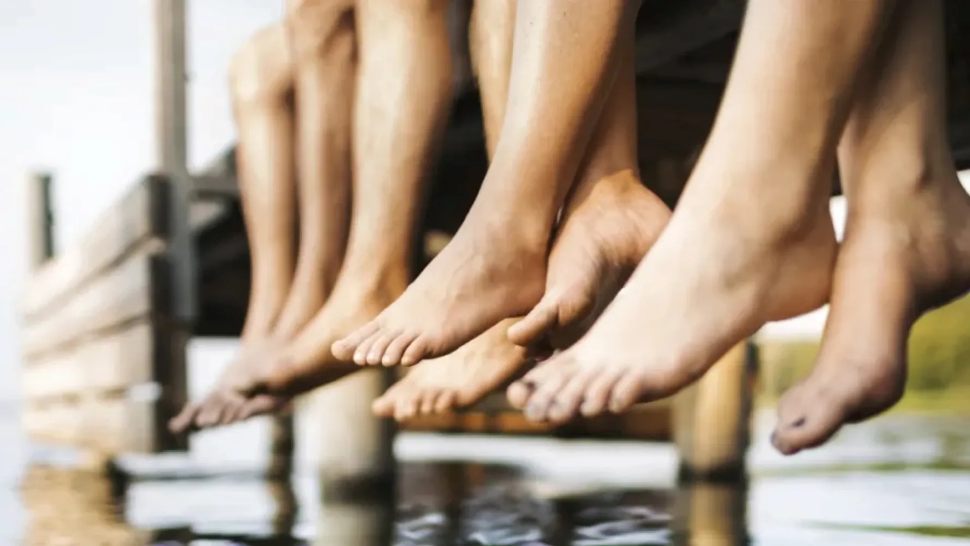 Group of people sitting on a wooden dock with feet dangling above calm water near greenery.