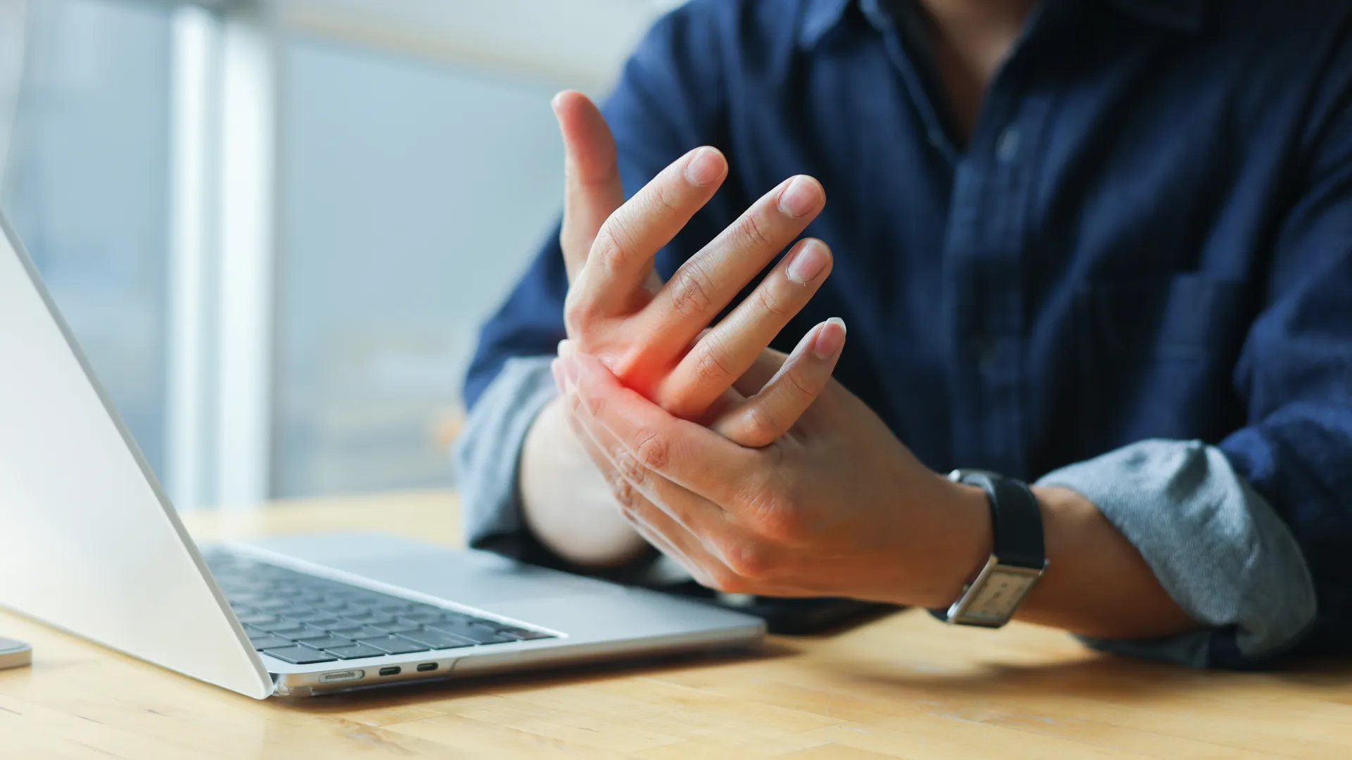 Person massaging wrist in pain while working on laptop at wooden desk indoors.