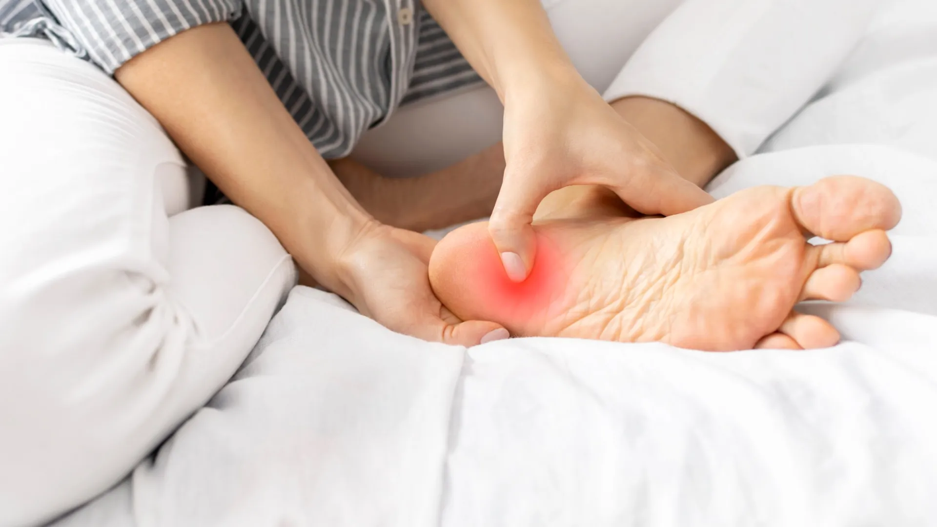 Person rubbing the sole of their foot highlighting pain area in red on a white bed.